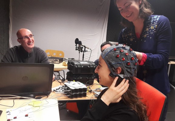 An EEG cap is being placed on the head of a subject by an experimenter on the right while another experimenter on the left is setting up the necessary software on the computer.